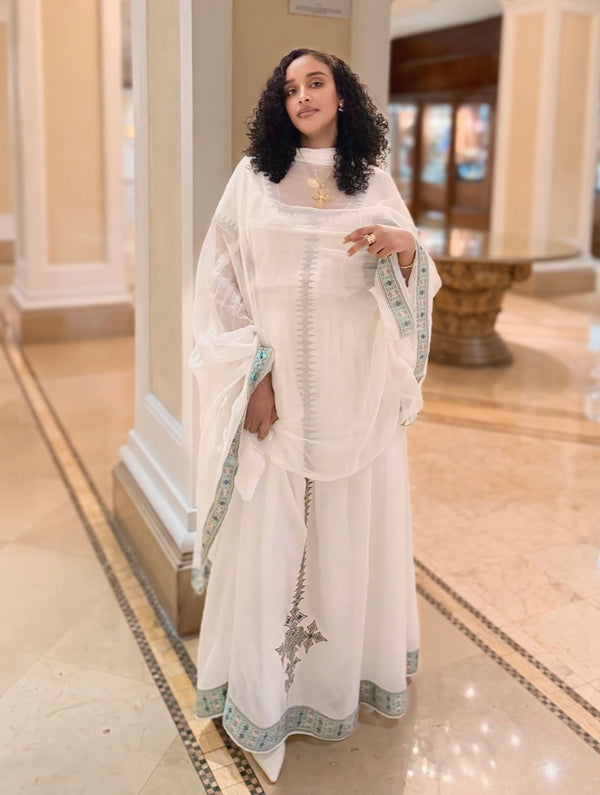 A woman wearing a traditional white Ethiopian dress with a matching sheer outer layer and light blue patterned borders