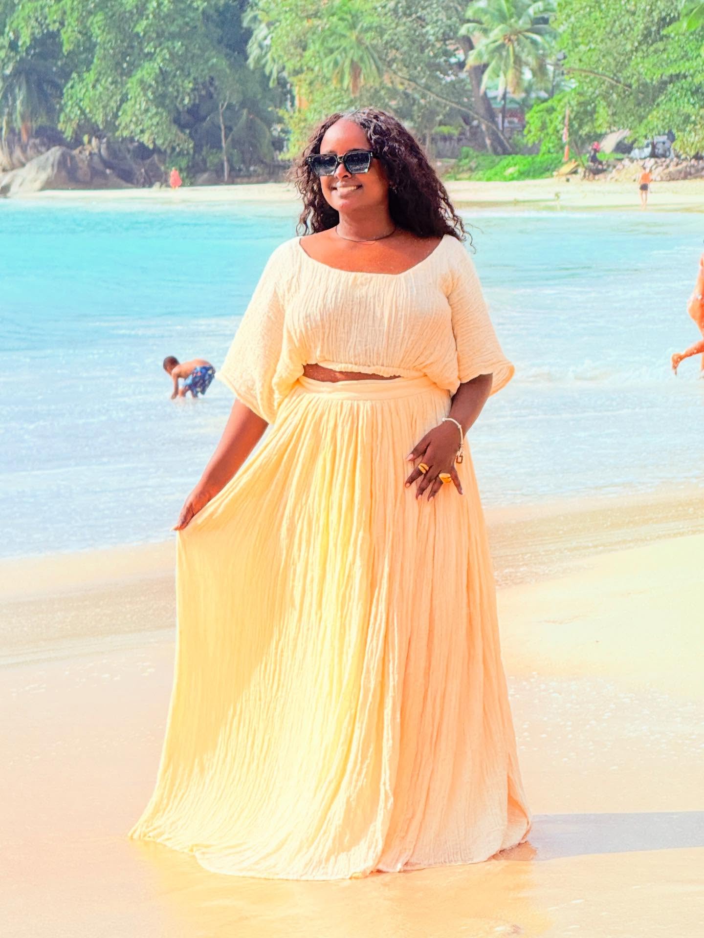 Woman in a off-white habesha dress standing on a beach with turquoise water and greenery in the background